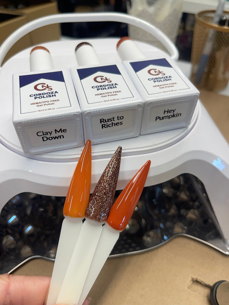 Three nail polish bottles with different colors held by a hand, with their packaging labeled 'Clay Me Down', 'Rust to Riches', and 'Hey Pumpkin' on a white surface.